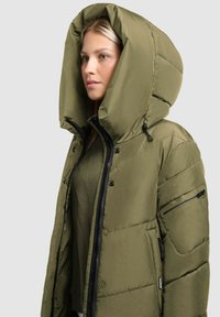 Chaqueta acolchada de color verde oliva con una gran capucha, que presenta un cierre de cremallera y bolsillos laterales. Textura suave con paneles cosidos y acentos en negro.