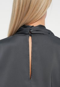 Blusa grigia scura con colletto alto, dotata di apertura a forma di chiave sul retro e due bottoni per la chiusura, realizzata in un tessuto liscio e morbido.
