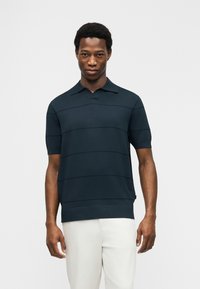 Άνδρας που φοράει σκουρόμπλε κοντομάνικο πουκάμισο με γιακά και διακριτικές οριζόντιες ρίγες και ανοιχτόχρωμο παντελόνι, στέκεται μπροστά σε απλό φόντο.