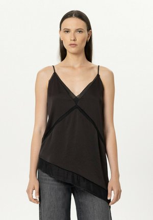 Young woman with straight brown hair wearing a black sleeveless V-neck top with thin straps and asymmetrical hem, paired with gray jeans.