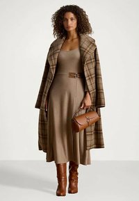 Woman with curly hair wearing a beige belted dress, brown plaid coat draped over shoulders, holding a brown handbag, and brown boots.