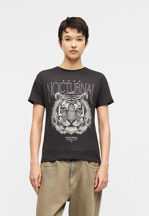Mladý človek s krátkymi tmavými vlasmi v čiernom tričku s detailnou tvárou tigra a nápisom "NOCTURNAL", v kombinácii s béžovými nohavicami.