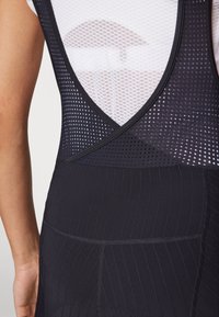 Svart atletisk tank topp med mesh-deler, V-formet rygg, og ribbet tekstur på den nedre delen. Merkbare sømdetaljer gjennom hele.