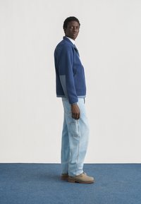 Νεαρός άνδρας που στέκεται πλάγια, φοράει μπλε φλις μπουφάν, ανοιχτό μπλε τζιν τύπου κάργκο και μπεζ μποτάκια σε μπλε πάτωμα.