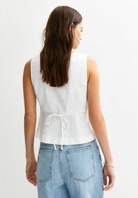 Haut sans manches blanc en lin, avec une taille cintrée et un détail noué dans le dos. Associé à un jean en denim bleu clair.