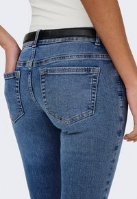 Gros plan d'une personne portant un jean bleu avec des poches arrière et une ceinture noire, main reposant dans la poche gauche.