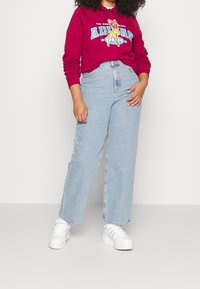 Woman wearing a red hoodie with a cartoon deer, light blue high-waisted jeans, and white sneakers standing against a plain background.