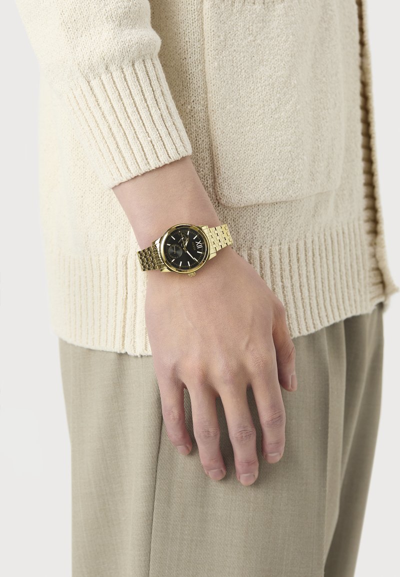 Main portant une montre en or avec un cadran noir, reposant sur le côté vêtu d'un pull en tricot crème et d'un pantalon beige.