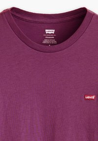 Purpurinė medvilninė marškinėliai su apvaliu kaklu, turintys raudoną Levi's logotipo emblemą kairėje pusėje ir banguotą kolorą.