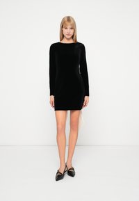 Vestido largo de terciopelo negro con mangas largas, escote en forma de barco, diseño ajustado y dobladillo asimétrico. Combinado con unos zapatos negros brillantes de punta afilada.