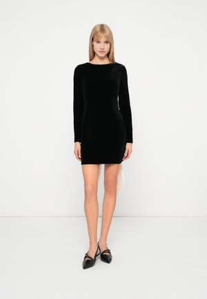 Vestido largo de terciopelo negro con mangas largas, escote en forma de barco, diseño ajustado y dobladillo asimétrico. Combinado con unos zapatos negros brillantes de punta afilada.