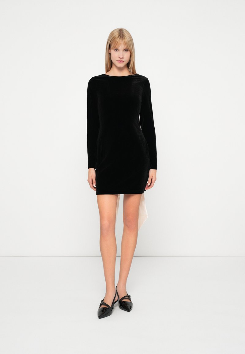 Vestido largo de terciopelo negro con mangas largas, escote en forma de barco, diseño ajustado y dobladillo asimétrico. Combinado con unos zapatos negros brillantes de punta afilada.