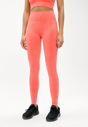 Vrouw die koraalkleurige legging met hoge taille en bijpassende cropped top draagt, staand met ontspannen handen langs het lichaam en zwarte sportschoenen.