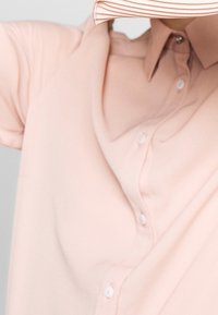 Chemise rose pâle à boutons avec col plié, présentant une texture lisse et de petits boutons blancs sur le devant.