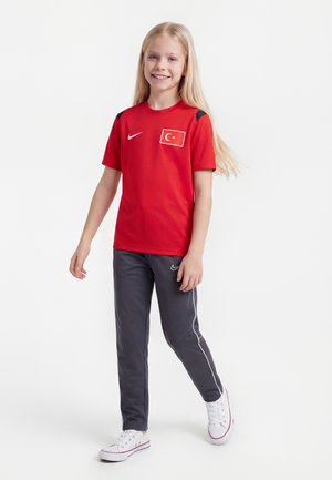 Młoda dziewczyna z długimi blond włosami chodzi uśmiechając się, ubrana w czerwoną sportową koszulkę z turecką flagą i czarne spodnie treningowe Nike.