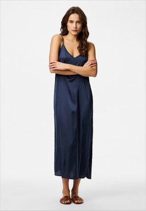 Kobieta z długimi brązowymi włosami stoi z założonymi ramionami, ubrana w granatową, bezrękawną sukienkę midi i brązowe płaskie sandały na białym tle.