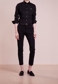 Person klädd i svart skjorta med knappar, svarta slim-fit byxor med bälte och svarta loafers, stående mot en blek rosa bakgrund.