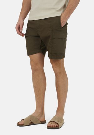 Man draagt donkergroene cargoshorts met zijzakken, een beige mouwloze top en beige instapsandalen tegen een witte achtergrond.