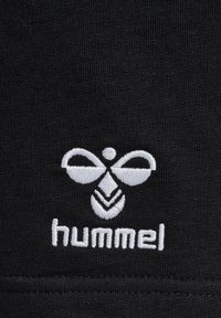 Balta siuvinėta stilizuota bitės logotipas su tekstu „hummel“ apačioje ant juodo tekstūruoto audinio fono.