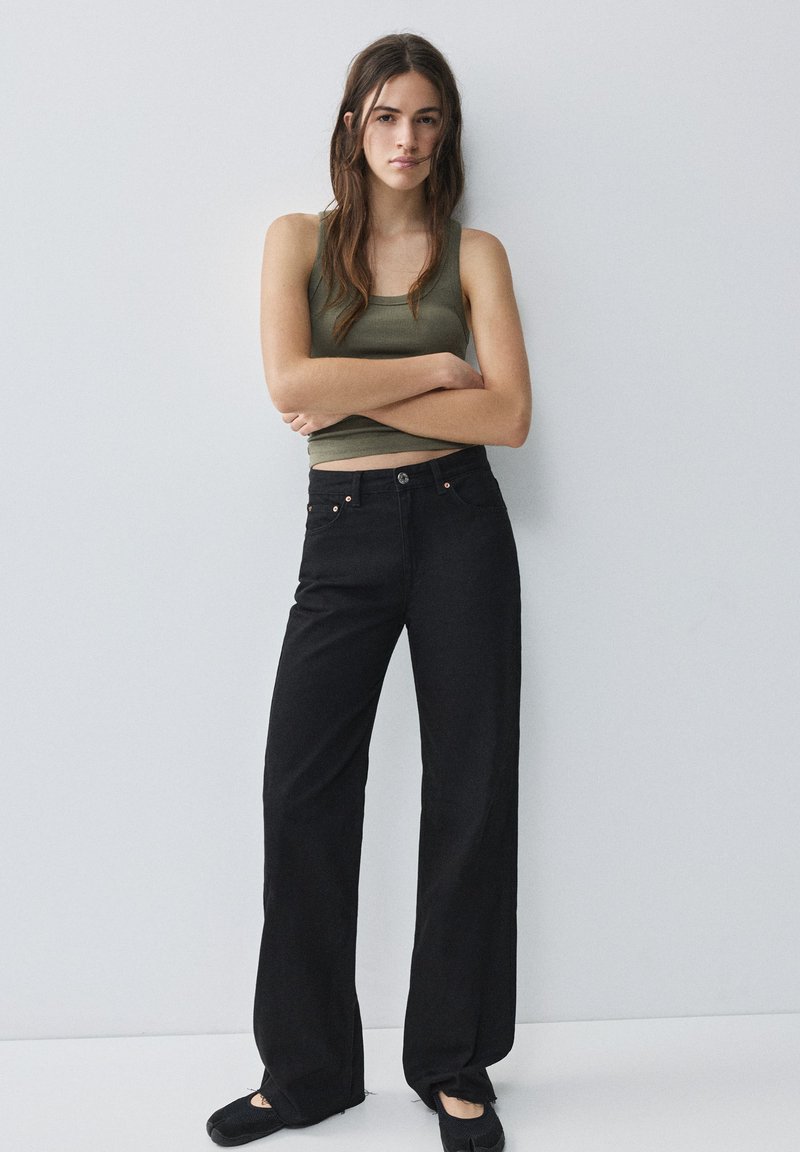 Jeune femme aux longs cheveux bruns se tient contre un mur blanc, portant un haut vert sans manches, un pantalon noir à jambes larges et des chaussures noires.
