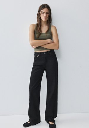 Jeune femme aux longs cheveux bruns se tient contre un mur blanc, portant un haut vert sans manches, un pantalon noir à jambes larges et des chaussures noires.