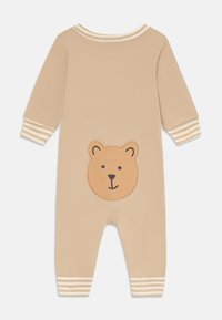 Romper beige para bebé con mangas largas, puños a rayas y un parche de cara de oso sonriente en la parte trasera cerca del fondo.