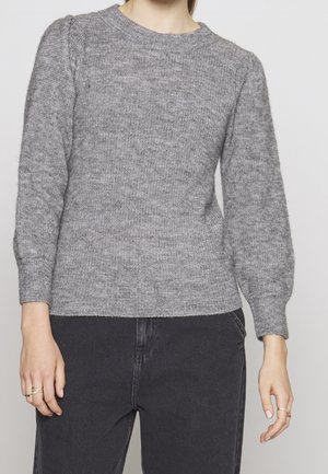 Femme portant un pull gris en maille avec des manches ballon et un pantalon noir taille haute, debout devant un fond neutre.