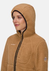 Beige fleecetröja med fastsydd huva, frontdragkedja och bröstficka. Svarta detaljer vid huva och ärmslut. Strukturerad yta.