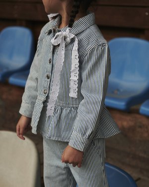 Παιδί με μπλε και λευκό κάθετα ριγωτό μπουφάν και παντελόνι με λευκό δαντελένιο φιόγκο, στέκεται κοντά σε μπλε πλαστικές θέσεις γηπέδου.