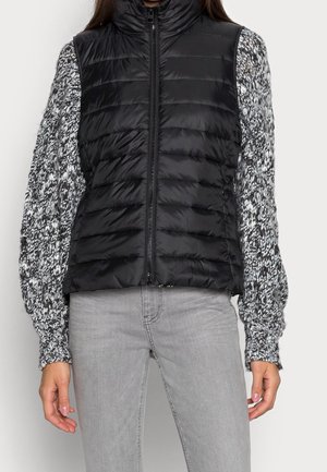 Persoon draagt een zwart doorgestikt vest met ritssluiting over een zwart-wit gebreide trui met lange mouwen en lichtgrijze spijkerbroek tegen een witte achtergrond.