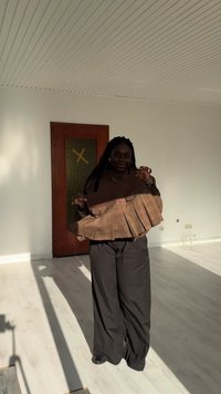 Kvinna som står i ett solbelyst rum och håller en brun rutig plisserad kjol med ribbat midjeband, iförd lösa mörka byxor och ett halsband.