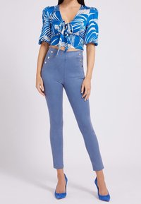 Top corto blu floreale con dettaglio annodato davanti, abbinato a jeans skinny azzurri a vita alta con bottoni laterali e scarpe con tacco blu.