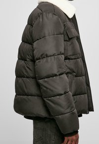 Pufjakke i mørkegrå med en hvid pelskrave, med et quiltet design, for- og baglommer samt lynlåslukning. Blød materialetekstur.