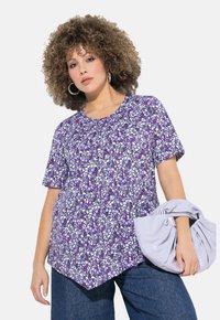 Top corto viola con motivo astratto bianco e lavanda, dotato di scollatura rotonda e orlo asimmetrico. La modella tiene una borsa di un delicato colore viola.