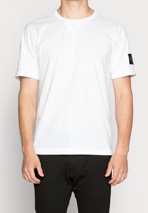 Wit katoenen T-shirt met korte mouwen, ronde hals en een logopatch op de linkermouw. Zachte textuur, standaard pasvorm.