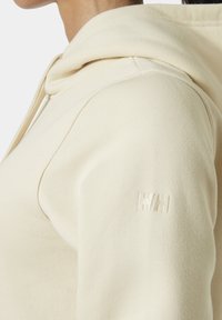 Beige hoodie met een soepele textuur, voorzien van een klein geborduurd logo op de linker mouw. Close-up van het schoudergebied.