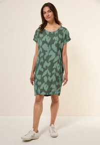 Vihreä kuvioitu mekko, jossa on väljä istuvuus, lyhyet hihat ja syvän kaula-aukon muoto. Yhdistetty valkoisiin tennareihin rentoa ilmettä varten.