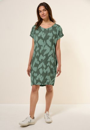 Grünes gemustertes Kleid mit lässiger Passform, kurzen Ärmeln und rundem Ausschnitt. Kombiniert mit weißen Turnschuhen für einen lässigen Look.