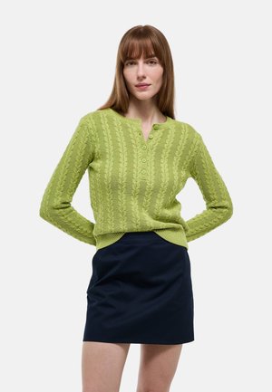 Femme aux longs cheveux roux portant un pull torsadé vert clair et une jupe bleu marine, debout, les mains dans le dos.