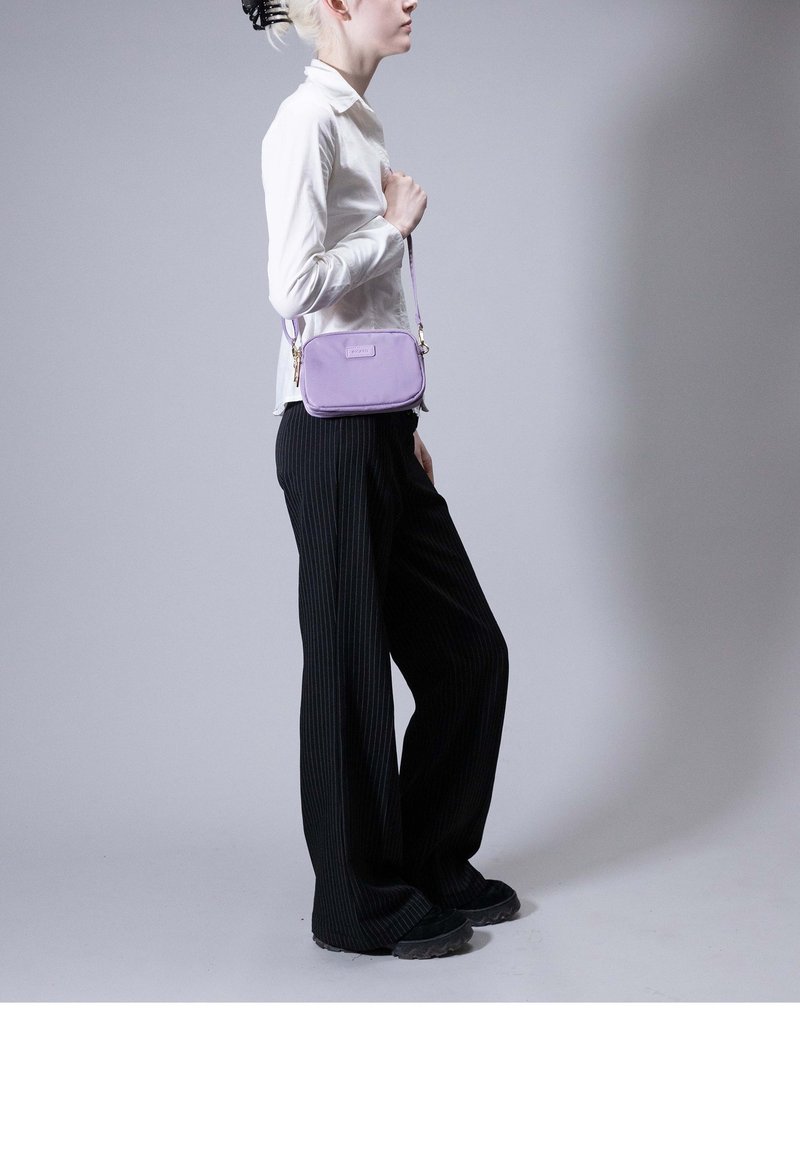 Persoon in wit overhemd en zwarte broek staat zijwaarts en houdt een kleine lavendelkleurige crossbodytas vast tegen een grijze achtergrond.