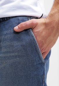 Džínové kalhoty ve středně modré barvě s mírným vyblednutím; rovný střih s rukou v kapse, která ukazuje šitý šev a jemné obvodové záhyby.