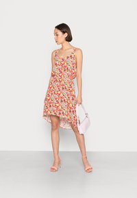 Vestido floral com um comprimento assimétrico em tecido multicolorido, apresentando um corte solto e alças ajustáveis. Acompanhado por uma bolsa rosa clara.