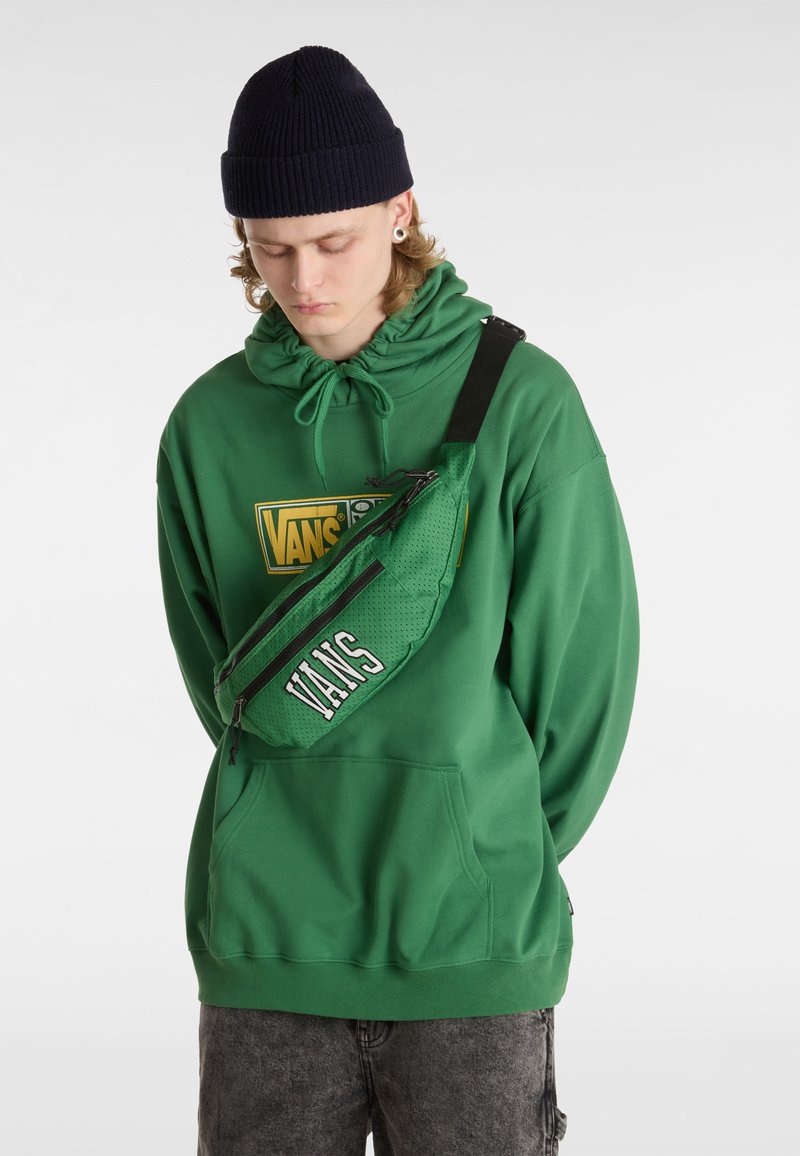 Felpa verde con tasca frontale e logo Vans; abbinata a una borsa a tracolla in rete verde con zip e testo Vans. Viene indossato un berretto lavorato a maglia scuro.