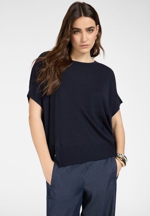 Donna con capelli castani lunghi che indossa una maglietta blu navy a maniche corte e pantaloni blu larghi, in piedi con le mani nelle tasche davanti a uno sfondo semplice.