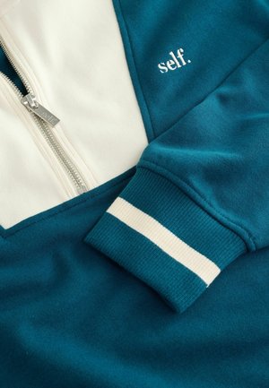 Türkise und cremefarbene Zip-Hoodie mit dem gestickten Wort "self." auf der Brust und gestreiften Bündchen. Nahaufnahme des Gewebes.