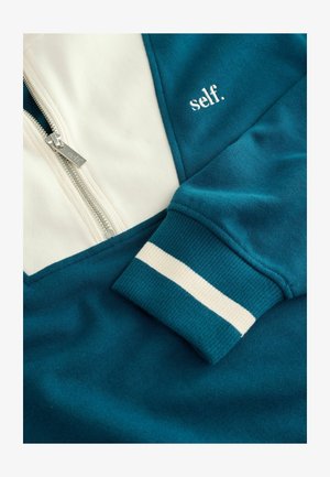 Sweatshirt zippé teal et crème avec "self." brodé sur la poitrine et des poignets rayés. Gros plan sur la texture du tissu.