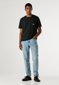 Μαύρο βαμβακερό t-shirt με τσέπη στο στήθος, φορεμένο με ανοιχτό μπλε τζιν και μαύρα παπούτσια. Το πουκάμισο έχει άνετη εφαρμογή και στρογγυλή λαιμόκοψη.