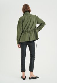 Green, lightweight jacket with a gathered waist, relaxed fit, and high collar, paired with black slim-fit pants and slip-on shoes.