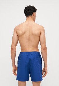 Blå svømmedrakt med elastisk liv, baklomme, laget av glatt stoff. Ingen synlige mønstre eller detaljer. Bakvinkel av modellen.
