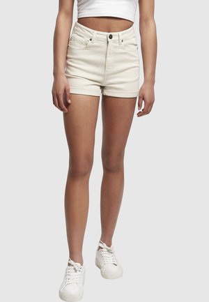 Kvinde iført højtaljede cremefarvede denimshorts, hvid croppet top og hvide sneakers, stående mod en ens grå baggrund.